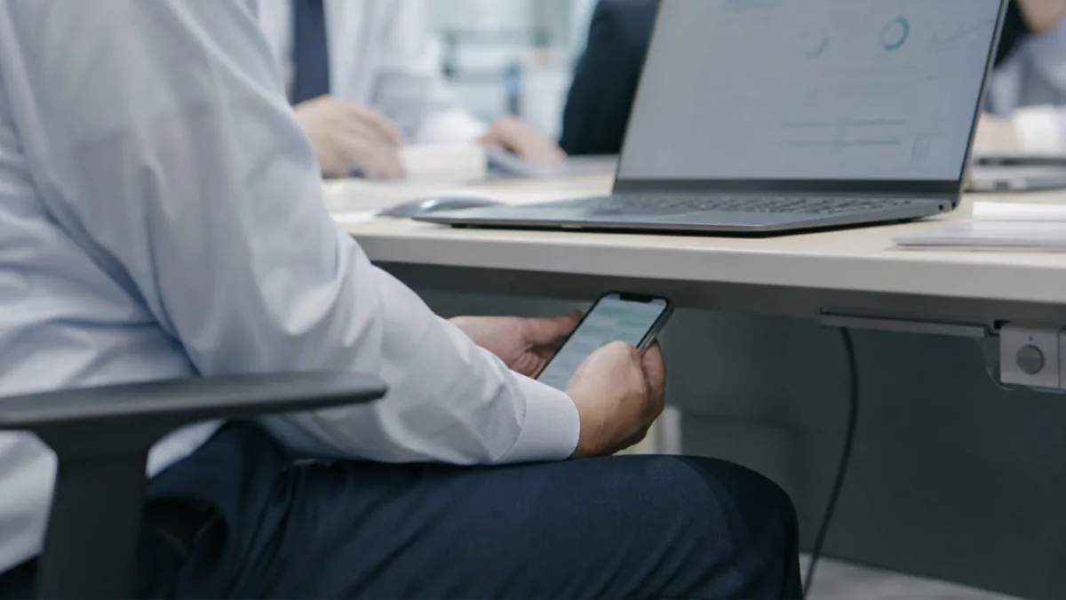 会社支給のノートPCが置かれたオフィスのデスクで、社員が机の下に隠すように自分のスマートフォンを操作している場面。生成AIの利用を禁止された社員が個人端末でこっそり使う「シャドーAI」の状態を表している